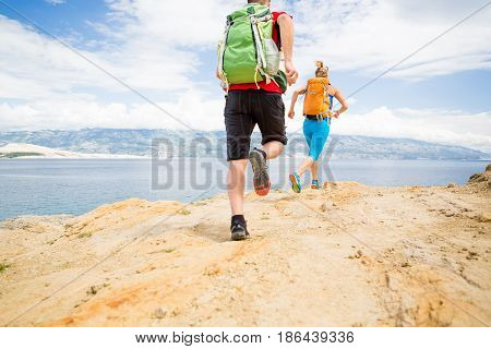 Couple runners running with backpacks on rocky trail at seaside and mountains. Young woman and man trail running on mountain path looking at beautiful inspirational landscape view.