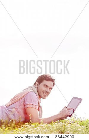 Side view portrait of young man using digital tablet while lying on grass against clear sky