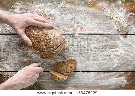 Baking background. Hands of baker cutting rye bread loaf with knife on rustic wooden table sprinkled with flour.