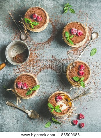 Homemade Italian dessert Tiramisu served in individual glasses with fresh mint leaves, raspberries and cocoa powder in sieve over grey concrete background, top view, copy space, vertical composition