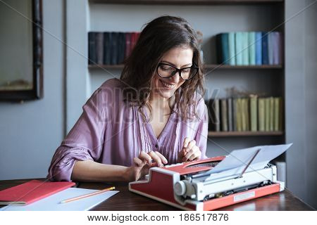 Portrait of a smiling mature authoress sitting at the table and using typewriter indoors