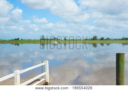 Green of sugar cane plantation across Clarence River with clouds reflected in calm water from jetty with yacht moored on other side near Australian town of Ulmara