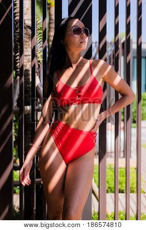 Sexy woman wearing red bikini swimsuit and sunglasses standing on the near swimming pool with artistic shadow pattern on body and face.