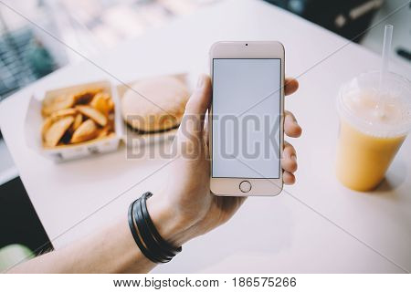 Young man's hand holding mobile phone. He is sitting in cafe and having his meal while searching for some information. Place for your advert or logo. Close up. Horizontal position of the phone