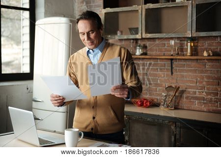 Correct mistakes. Positive father keeping smile on face bowing head while looking downwards at sheet of papers