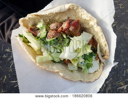 Hand holding traditional greek food souvlaki also known as gyros with meat in pita bread and white paper bag served at a food festival.