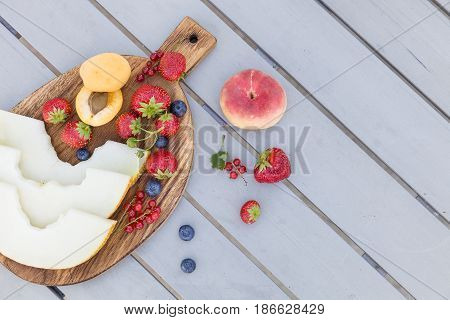 Top view on wooden board with fresh organic fruit and berries: melon strawberries blueberries apricot peach and red current on grey background. Healthy snack