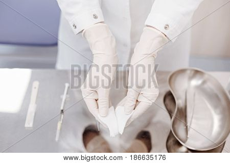 Everything is sterile. Close up of a nice professional doctor wearing disposable gloves and holding pieces of bandage while standing over the table