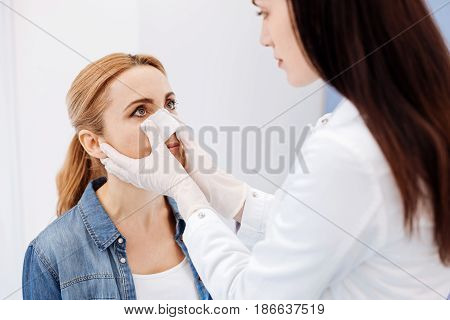 Nose reshaping. Nice female pleasant doctor standing in front of her patient and putting a medical dressing on her nose while preparing to do a nose surgery