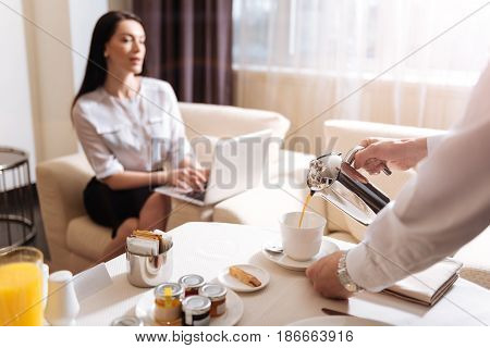 Morning coffee. Professional nice male waiter holding the coffee pot and pouring a drink into the cup while serving breakfast