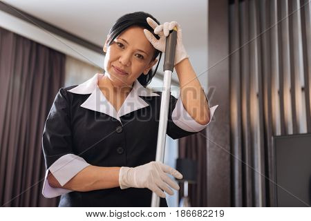 Hard working woman. Positive professional delighted chambermaid holding a mop and looking at you while cleaning the hotel room
