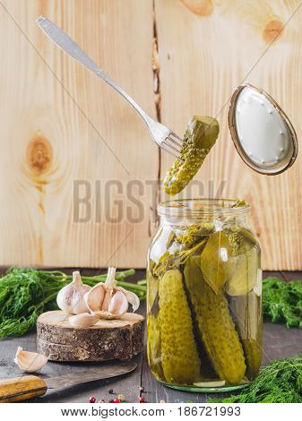 The fork holds a pickled cucumber that just has been taken from glass jar. Pickled cucumbers on wooden background. Pickles in glass jar with garlic, peppercorn and fresh dill. Flying food.