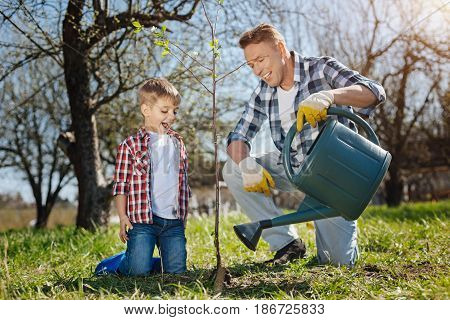 Taking care of nature. Happy father watering a newly planted tree with his adorable son while both standing on knees in a sunny spring day
