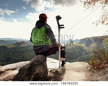 Tired Tourist With Medicine Crutch  And Broken Leg Fixed In Immobilizer Resting On Summit.