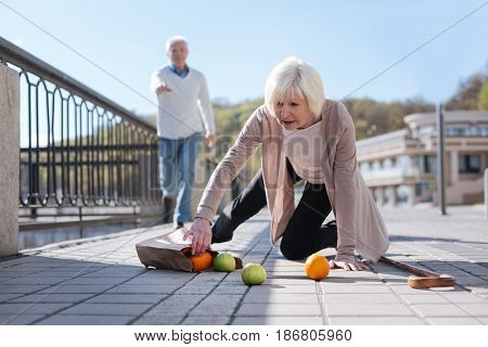 Weakness reason for troubles. Pleasant grey headed sad lady kneeling on the ground clamping greengrocery while middle-aged man going to her