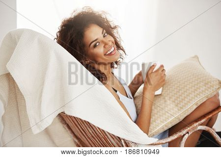 Beautiful african girl in sleepwear smiling looking at camera holding cup sitting relaxing in chair at home.