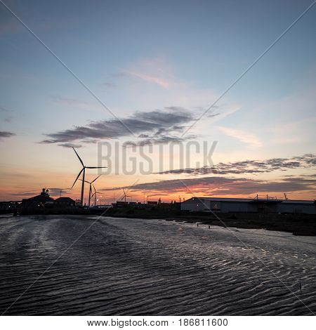 Dusk sunset view of the Port of Tilbury with the low tide banks of the River Thames and wind turbines turning in the background.