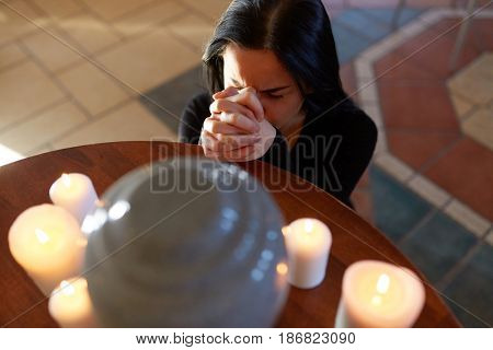 people and mourning concept - sad woman with cinerary urn and candles praying at funeral in church