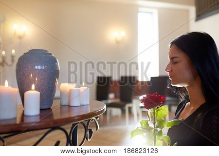 people and mourning concept - sad woman with red rose and cinerary urn at funeral in church