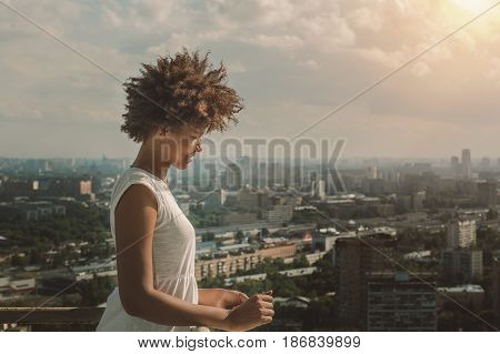 Young charming black Brazilian teen girl with beautiful curly afro hair in white dress standing on observation point very high and smiling sunny summer cityscape below in blurred background