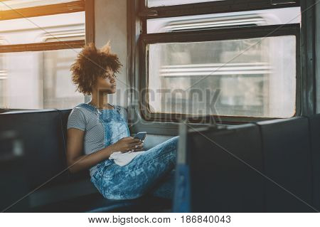 aCharming pensive black teenage female sitting inside carriage of suburban electric train and using smart phone attractive young mixed girl with cell telephone going by train and looking in window