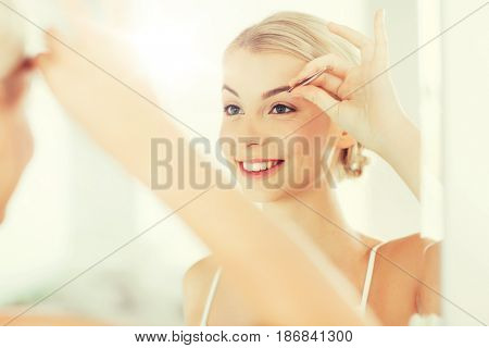 beauty and people concept - smiling young woman with tweezers tweezing eyebrow and looking to mirror at home bathroom