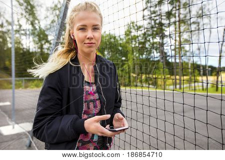 Portrait of beautiful fit woman holding mobile phone while listening music by fence