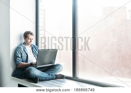 handsome hipster modern man designer working home using laptop at home siting on windowsill