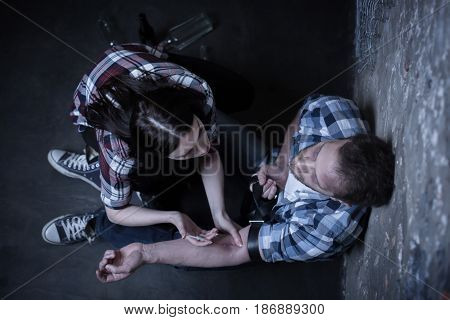 Trying to become happy. Lonely stoned weak couple sitting on the ground in the darkness while sharing drug dose and making syringe injection of heroin
