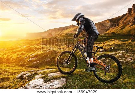 Ryder in full protective equipment on the mtb of two pendant bicycles rides along the green grass towards the sunset in the rays of the sunset sun against the backdrop of a landscape of rocky terrain under the setting sun and clouds.