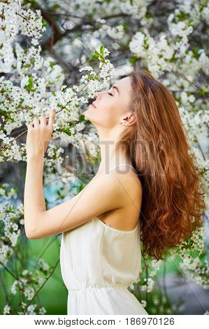 Young Beautiful Woman In Circlet Of Flowers Enjoying Smell Of Blooming Tree On A Sunny Day. Close Ey