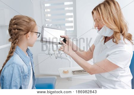 Look how we will do it. Dedicated neat prominent doctor giving her patient a lecture on dental health and showing some examples on her gadget while running a consultation
