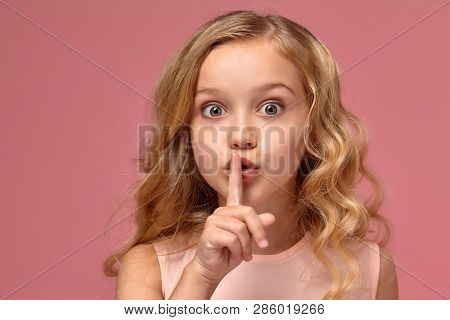 Little Girl With A Blond Curly Hair, In A Pink Dress Is Posing For The Camera