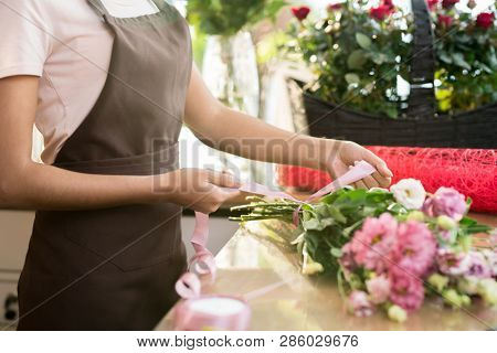 Florist in apron tying nude pink silk ribbon aroung fresh floral bouquet consisting of various flowers