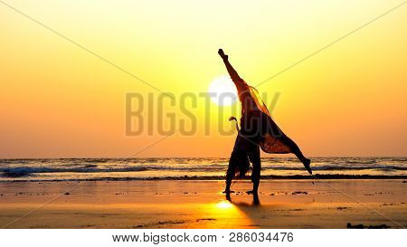 Silhouette Of Young Gymnast Woman Doing Handspring On Sandy Beach At Sunset