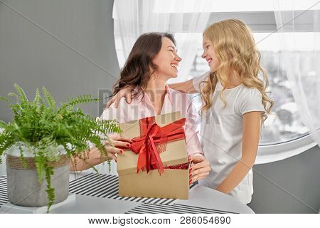Cheerful Daughter Hugging Happy Mother. Beautiful Brunette Opening Box With Red Bow On White Table W