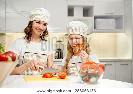 Beautiful Woman And Her Daughter Cooking Together Fresh Vegetable Salad. Cute Girl Biting Pepper Sli