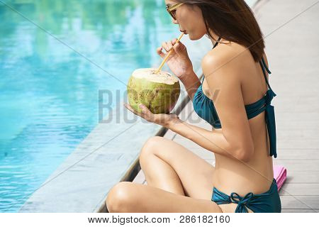 Beautiful Woman In Bikini Resting At Poolside And Drinking Coconut Drink From Straw Outdoors At Reso
