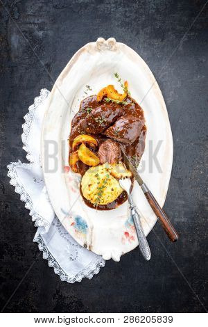 Traditional German braised pork cheeks in brown red wine sauce with fried mushed potatoes and pumpkin as top view on a shabby chic design white plate with copy space 