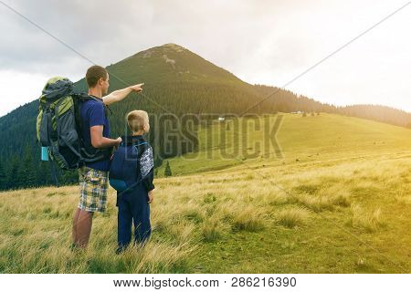 Father And Son With Backpacks Hiking Together In Summer Mountains. Back View Of Dad And Child Holdin
