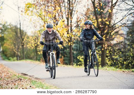 A Senior Couple With Electrobikes Cycling Outdoors On A Road In Park In Autumn.