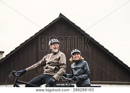 Active Senior Couple With Electrobikes Standing Outdoors In Front Of A House.