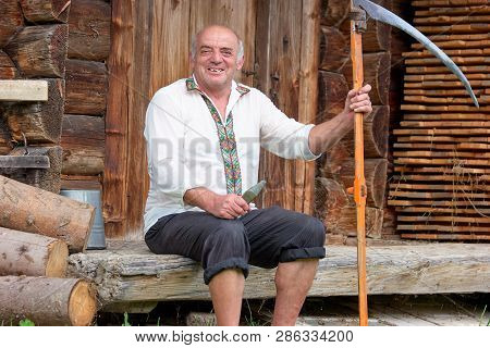 Senior Man In Hutsul Clothes With Scythe. Old Man In Embroidered Shirt Sitting In Front Of His Old W