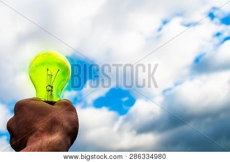 Green Bulb In Hand On Blue Sky Background. Hand Holding Light Bulb On A Background Sky And Sun. The 