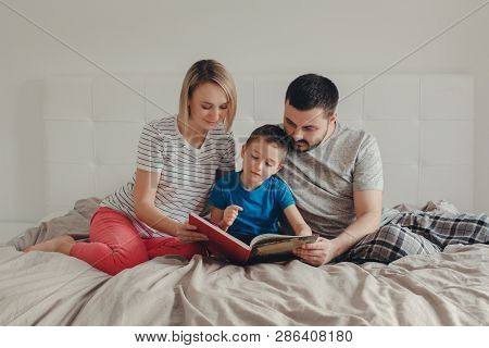 Family Of Three People Sitting On Bed In Bedroom Reading Book. Mother, Father And Boy Son At Home Sp