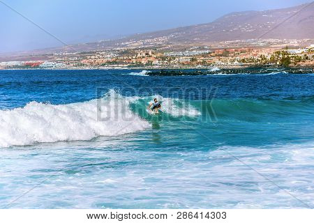 Costa Adeje, Spain - February 8, 2019: Surfer Girl On A Ocean Wave Against The Popular Resort In The
