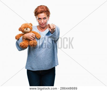 Senior caucasian woman holding teddy bear over isolated background pointing with finger to the camera and to you, hand sign, positive and confident gesture from the front