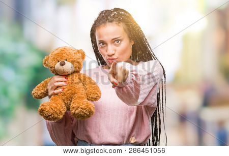 Young african american girl holding teddy bear over isolated background pointing with finger to the camera and to you, hand sign, positive and confident gesture from the front
