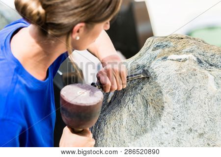 Female stonemason working on boulder with sledgehammer and iron