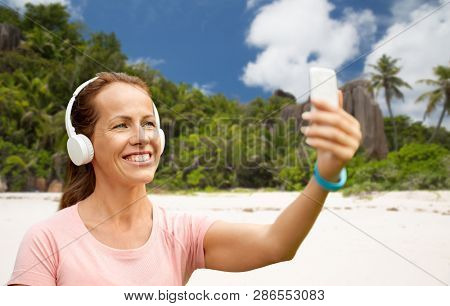 fitness, technology and healthy lifestyle concept - smiling woman in headphones listening to music and taking selfie by smartphone over tropical beach on seychelles island background